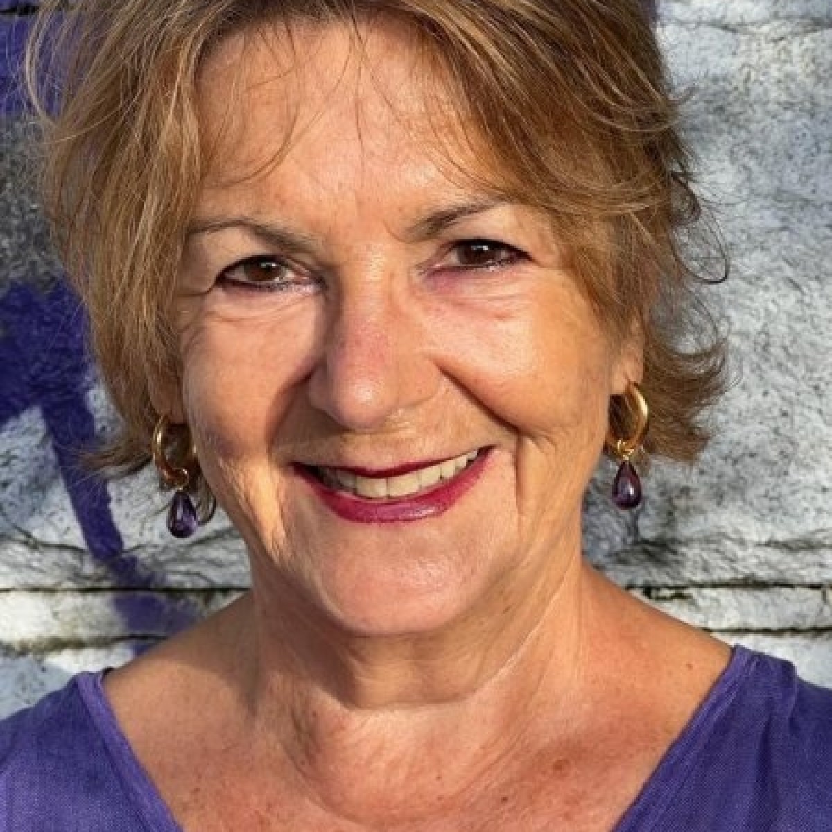 Smiling woman with short hair and purple earrings standing against a stone wall.