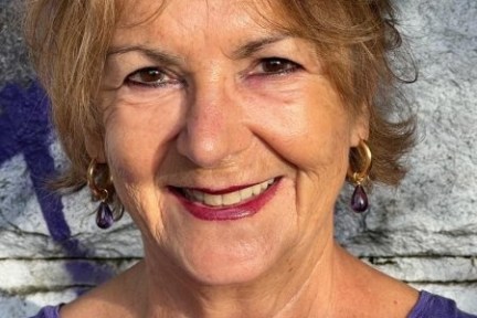 Smiling woman with short hair and purple earrings standing against a stone wall.