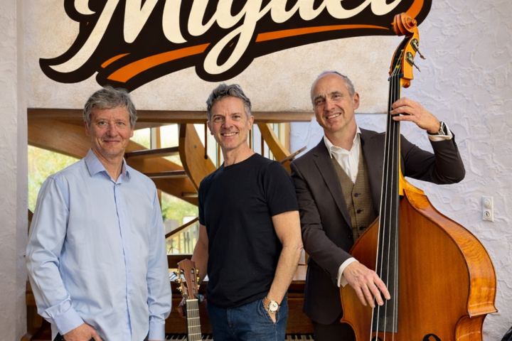 Three men with musical instruments standing under 'de Miguel' sign.
