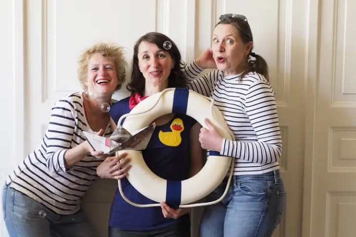 Three women with a lifebuoy and toy boat, smiling indoors.